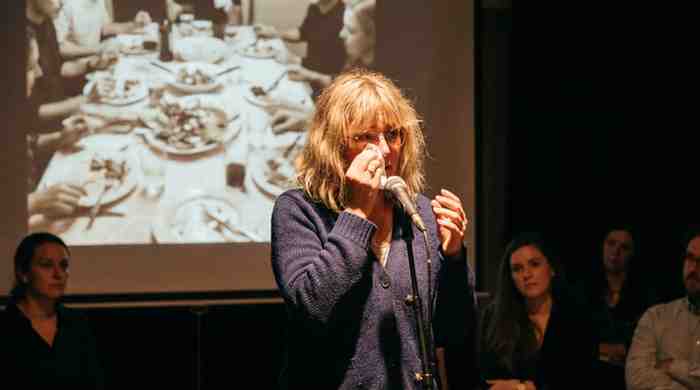 A woman with glasses and a navy cardigan stands at a microphone, wiping her eyes during a presentation while a faint black-and-white photo of a busy dinner table is projected behind her.