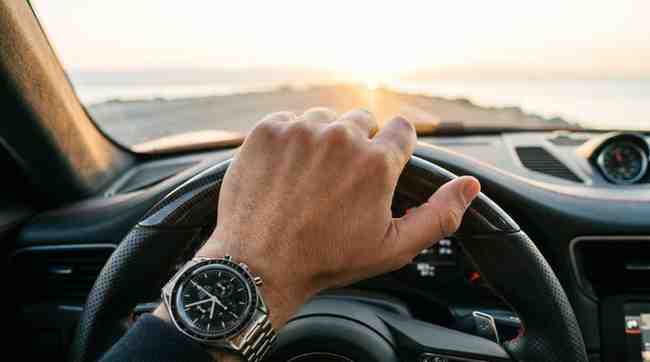 First-person view from inside a modern car showing a professional's hands in leather driving gloves gripping the steering wheel on a highway heading straight into a bright golden sunrise.