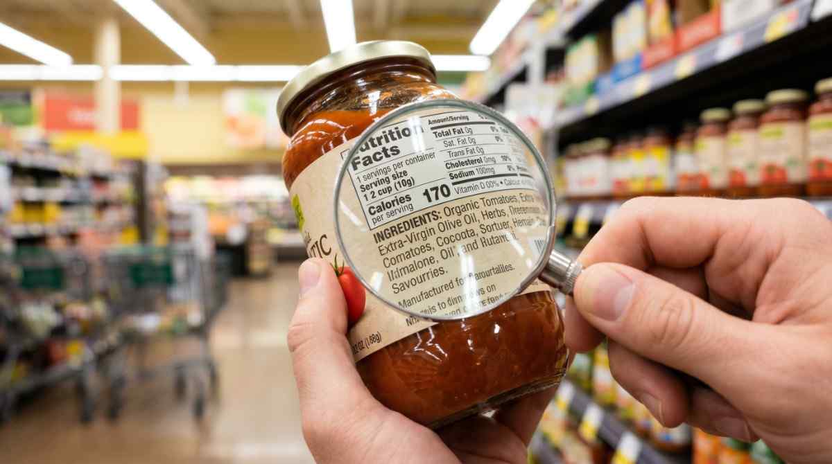 Close-up of a person's hands holding a food jar in a supermarket, focusing on the nutrition facts label showing calories, ingredients like organic tomatoes, and extra-virgin olive oil.https://www.devchandrika.com/