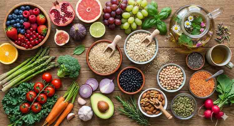 A colorful flat lay of plant-based healthy foods featuring fresh fruits, green vegetables, grains, legumes, nuts, seeds, and a glass teapot on a wooden background from devchandrika.com.